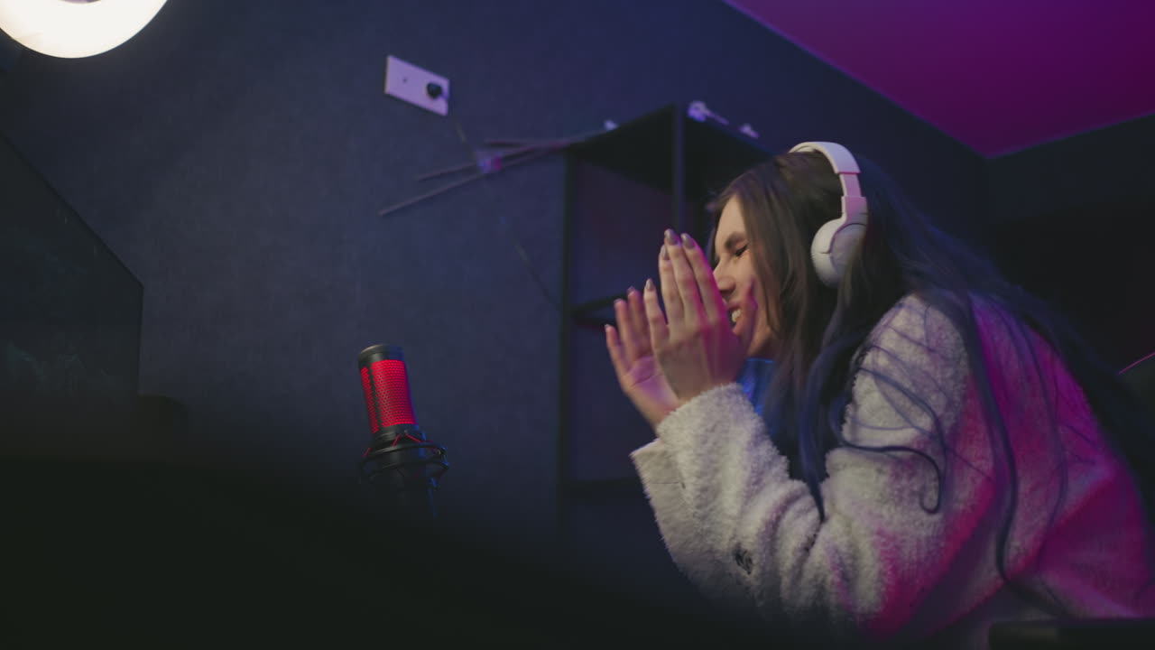 joyful young gamer wearing white headset and cozy sweater leans forward to fix her hair after winning computer game, seated at desk with microphone in dimly lit neon-accented gaming studio
