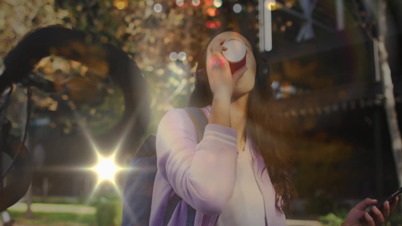 Female student drinking disposable cup holding smartphone on sunny campus with floating data icons