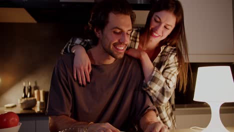 una chica morena feliz con una camiseta naranja y una camisa a cuadros se acerca a su novio por detrás y lo abraza. un chico moreno feliz leyendo un libro y abrazando a su novia morena en un apartamento moderno en la cocina por la noche