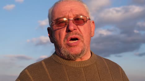 portrait of an elderly man with glasses and a beard gesturing and talking outdoors at sunset