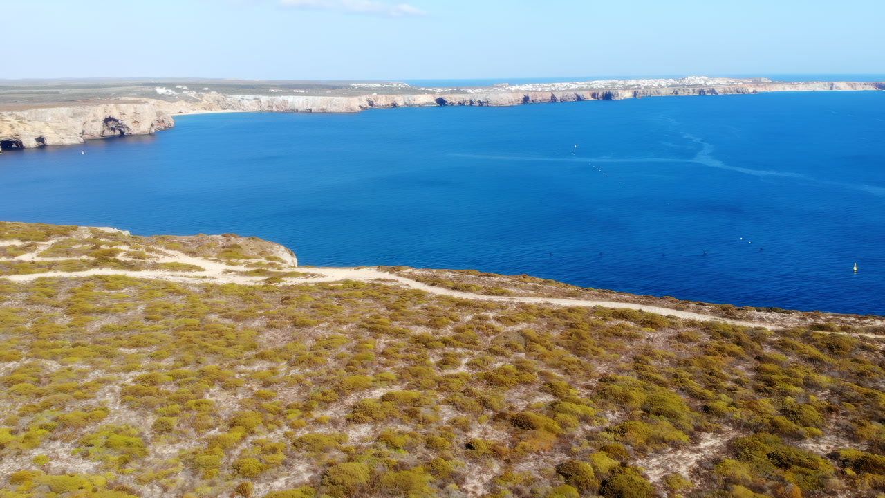 Aerial Cliff Path Overlooking Azure Bay, Sunlit Scrubland And Winding Trail Along Peaceful Coastline. Media