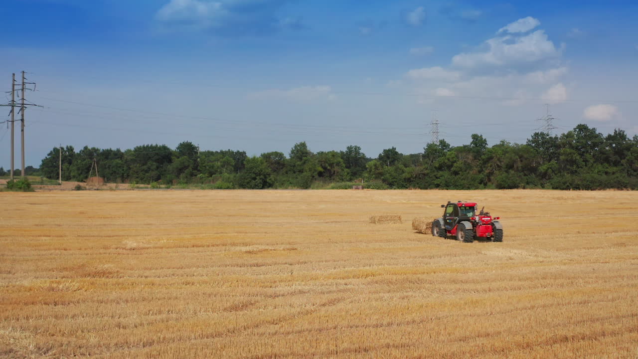 Approaching little red excavator piling the hay bales. Agricultural machinery working in the field after harvesting crops.