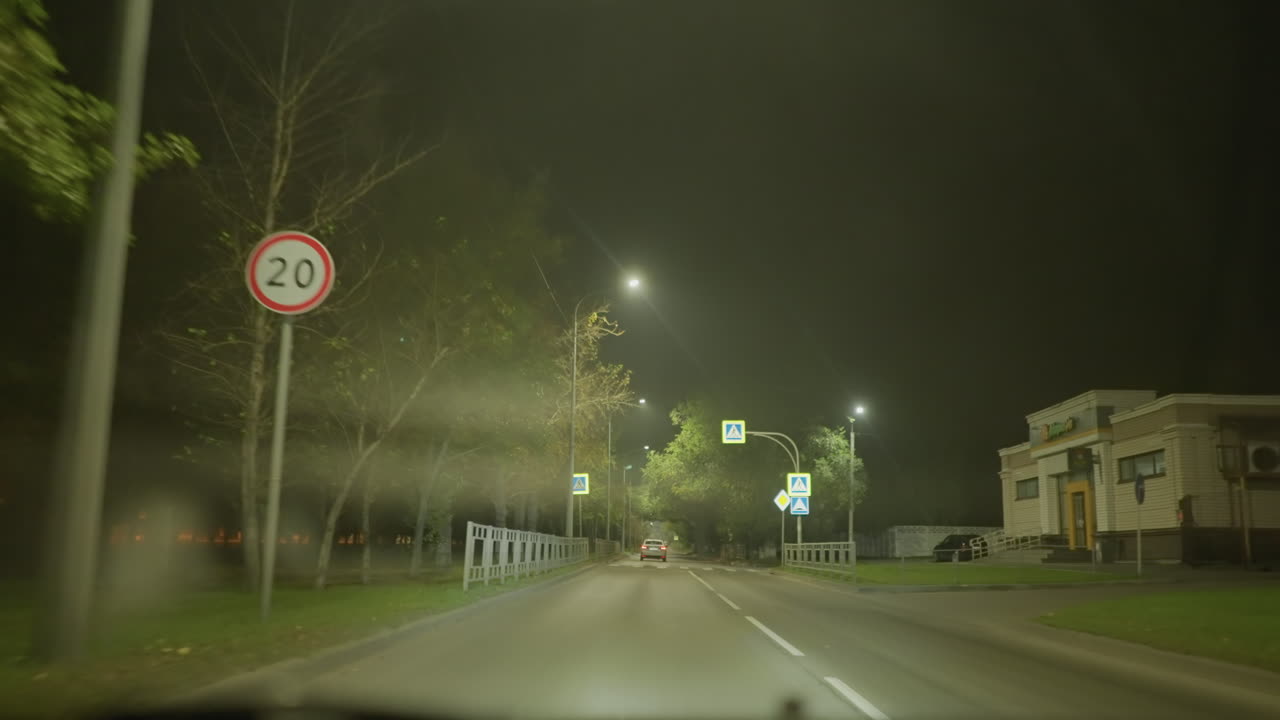 조용한 교외 지역의 밤거리를 운전하는 모습이에요. 운전석 시점에서 헤드라이트가 안개와 눈부심을 가르며 나아가고, 가로등이 은은하게 빛나고 있어요. 도로 표지판과 횡단보도가 보이고, 차선은 비어 있습니다.