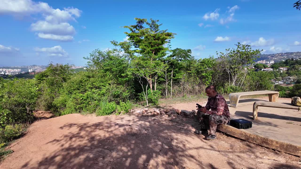 Uomo latino adulto siede con un telecomando pilotando un drone. Natura verde e soleggiata.