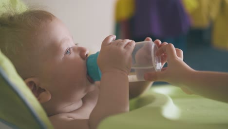 child-drinks-water-with-sister-help-sitting-in-highchair