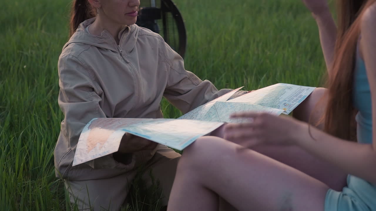 woman in beige flight suit seated on grass holding unfolded map while couple points at map beside fan cage and deflated colorful balloon fabric on farmland