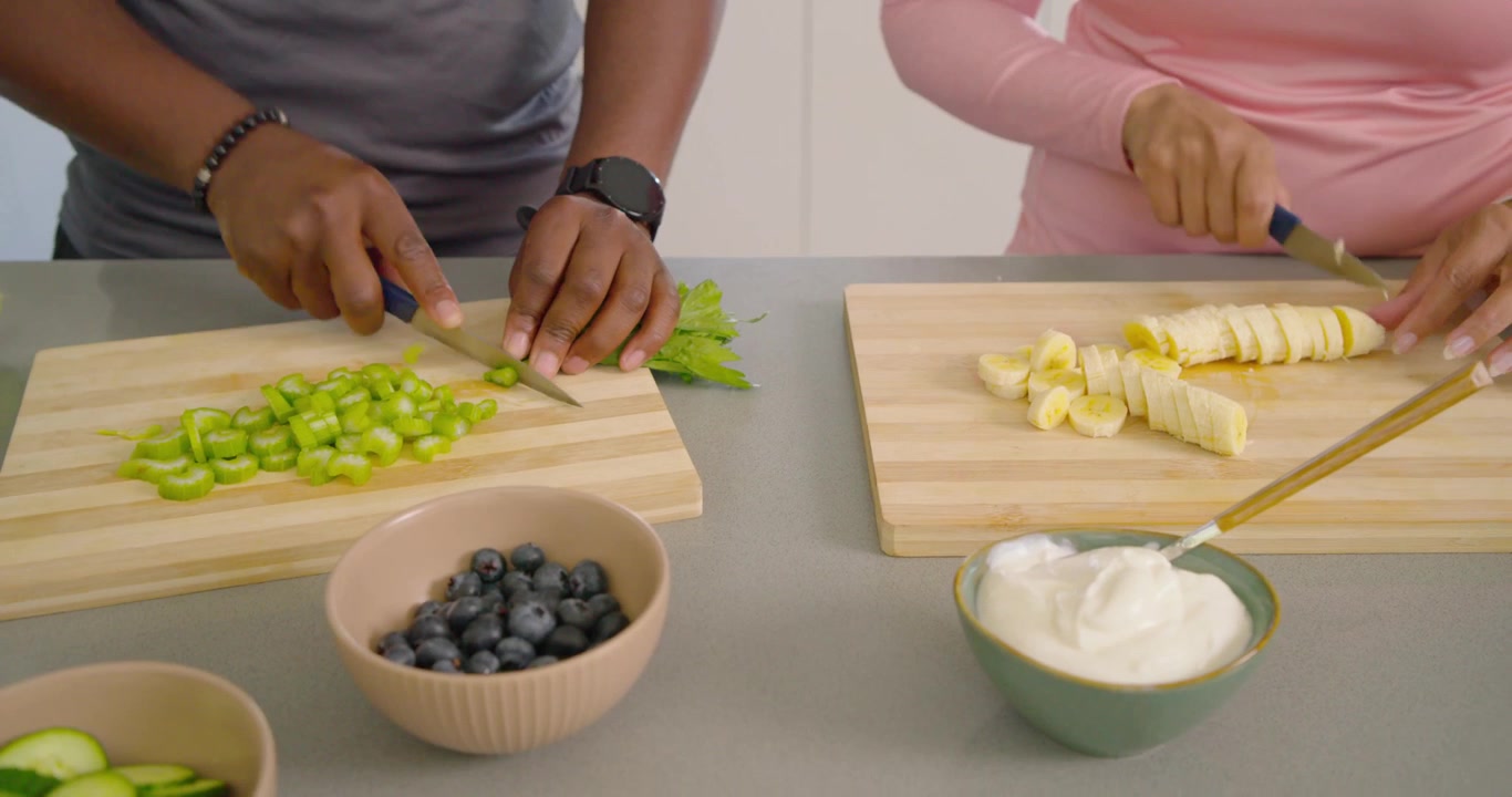 Coppia afroamericana che taglia il sedano e affetta la banana sul bancone, preparando ciotole di yogurt su taglieri.