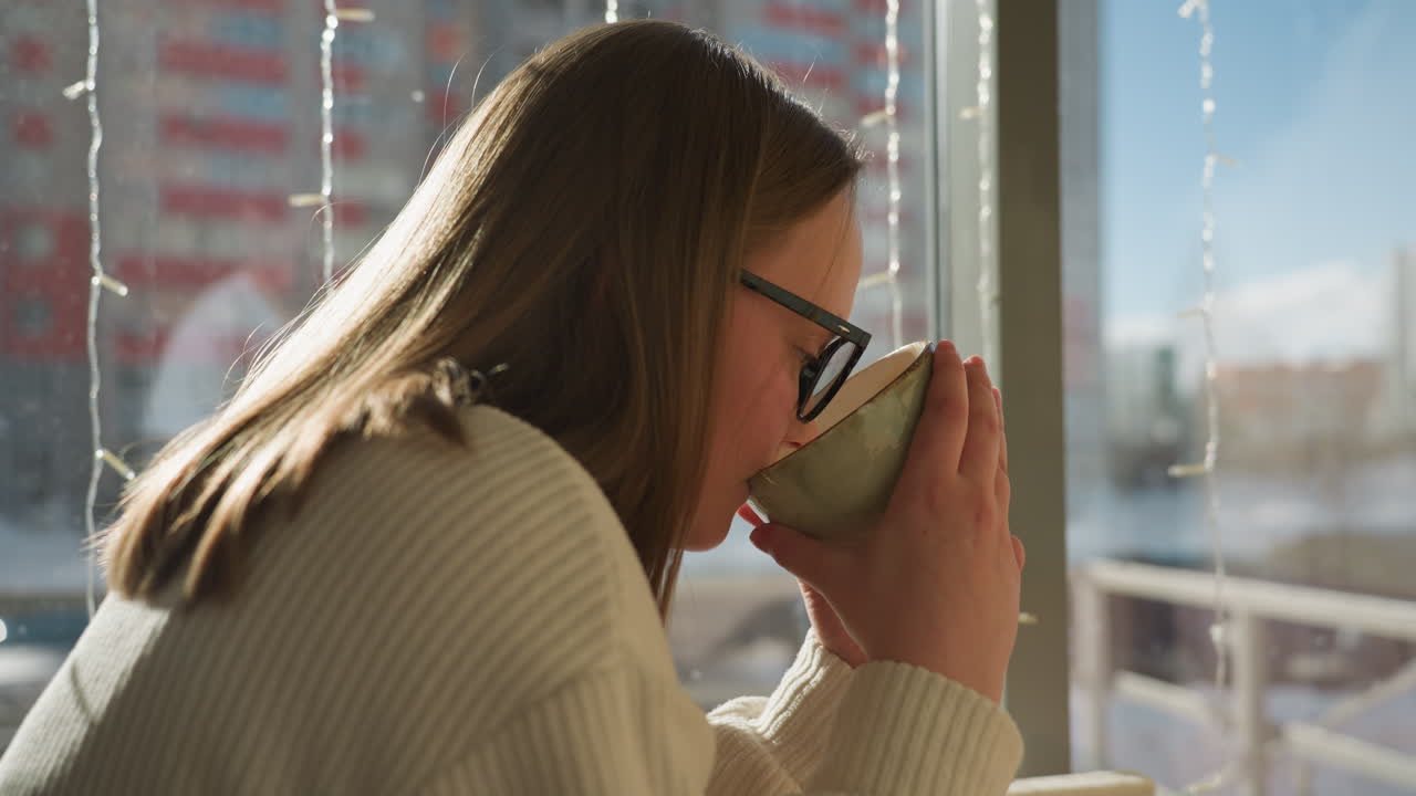 Side view of lady in cream sweater sipping coffee beside glass panel, sunlight pouring in, decorative lights hanging, snowy cityscape with parked cars and high rise buildings in background