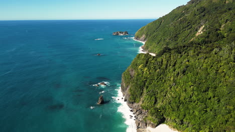 Aerial-shot-of-the-waves-crashing-into-the-thick-forest-covered-cliffs-of-the-west-coast-of-New-Zealand-on-a-sunny-day,-orbiting-shot