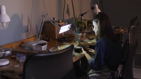 focused caucasian female jeweller sitting at desk, making jewelry in workshop