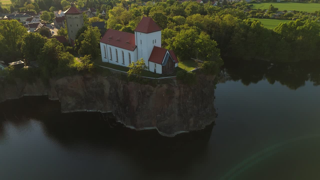 Betagende luftfoto af Wehrkirche Beucha, der ligger på en høj klippe over et oversvømmet stenbrud. Den middelalderlige kirke gløder i det varme solnedgangslys, omgivet af skov og rolige vande