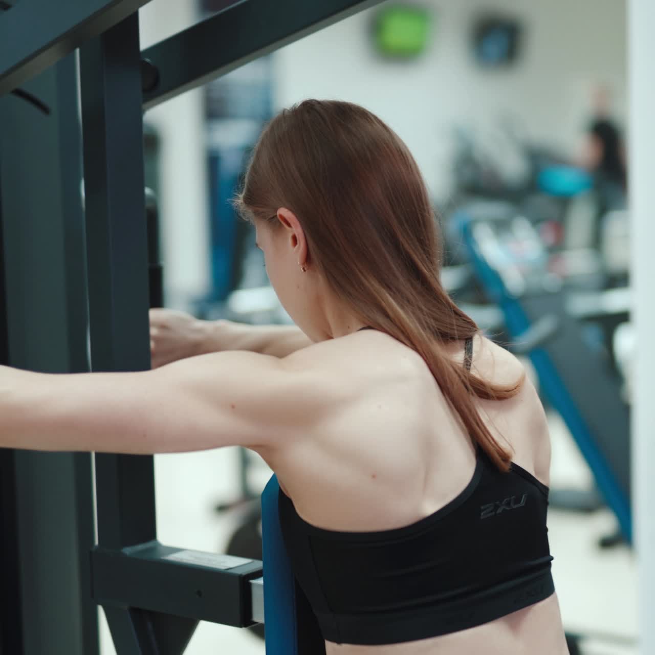 A strong young woman performs an exercise on the simulator for back and triceps. Blurred Background. Camera motion to right. Close-up