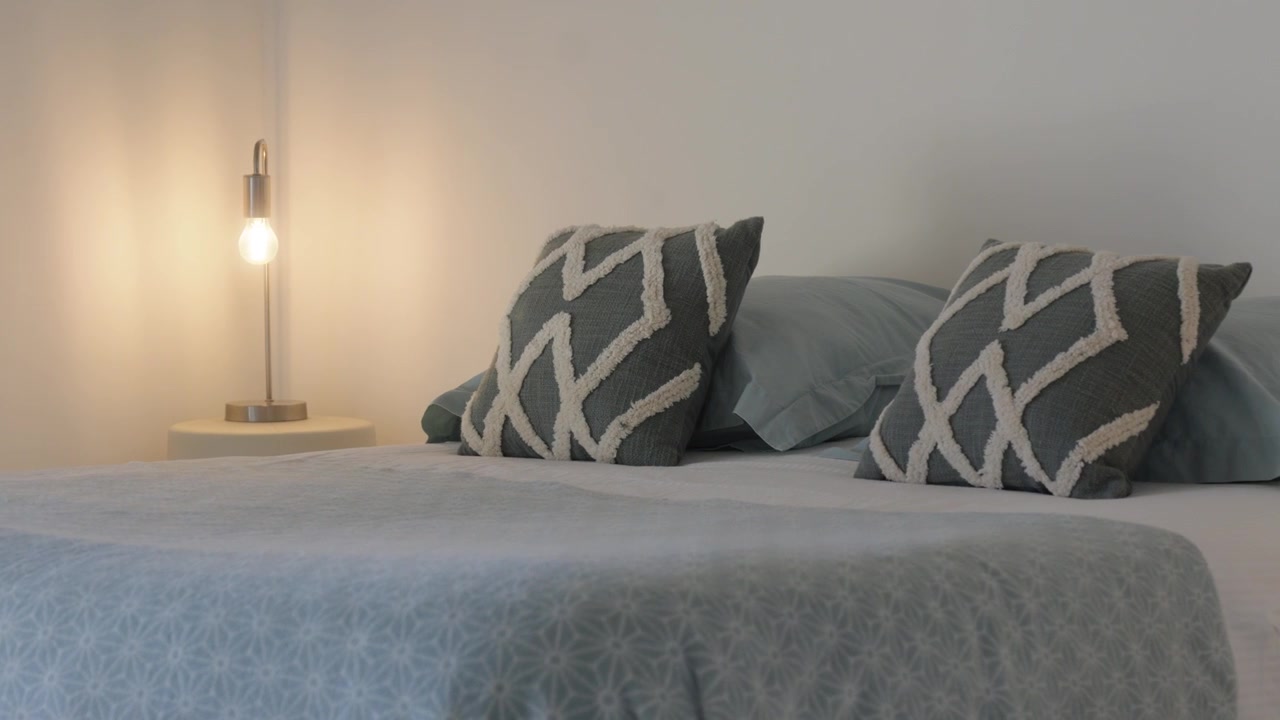 La telecamera inquadra un accogliente letto in camera da letto con cuscini decorativi e una lampada accesa.