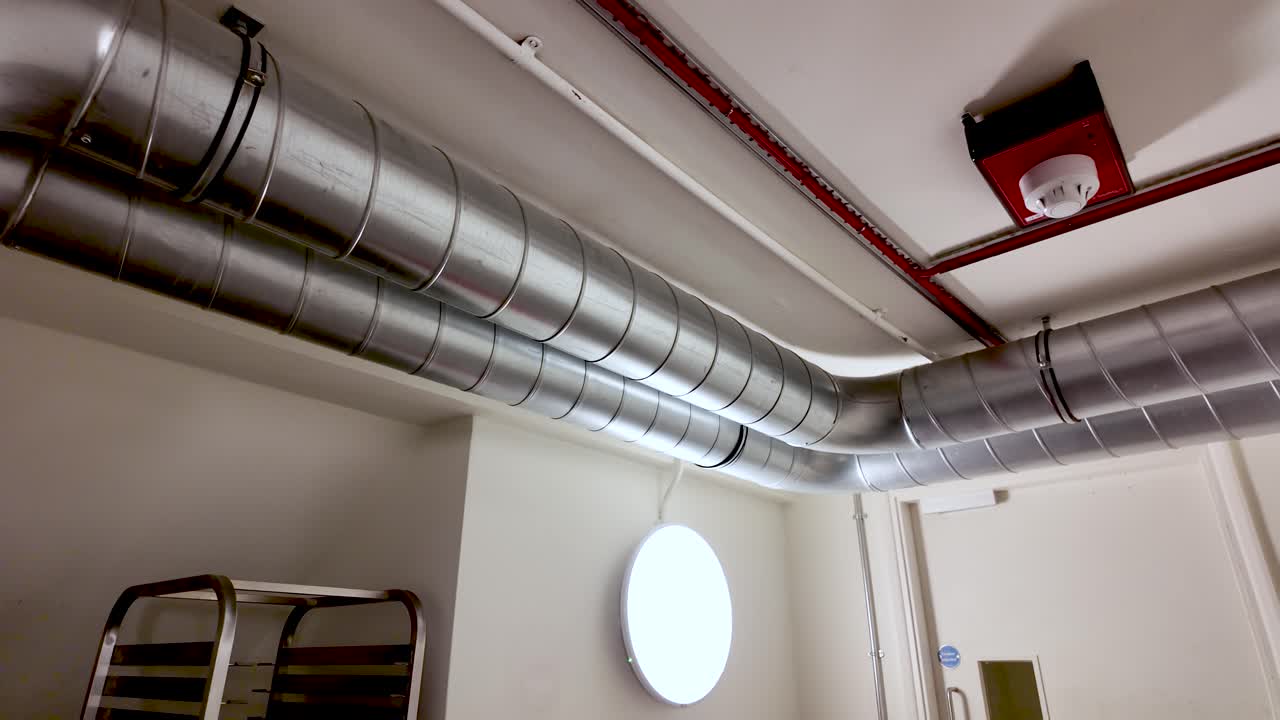 Un sistema di condotte metalliche corre lungo un soffitto bianco, trasportando l'aria attraverso uno spazio industriale e funzionale.
