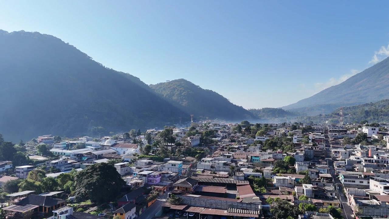 Veduta aerea con drone su San Lucas, Guatemala, cattura quartieri tropicali, nuvole luminose, palme e una catena montuosa vulcanica circostante con cime lussureggianti e boscose, creando un paesaggio vibrante