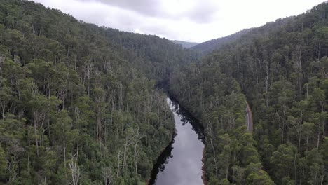 Slow-aerial-drone-pan-up-over-crystal-clear-still-river-stream-running-through-forest-trees-with-mountain-range-in-background-and-road-alongside-on-moody-clear-day