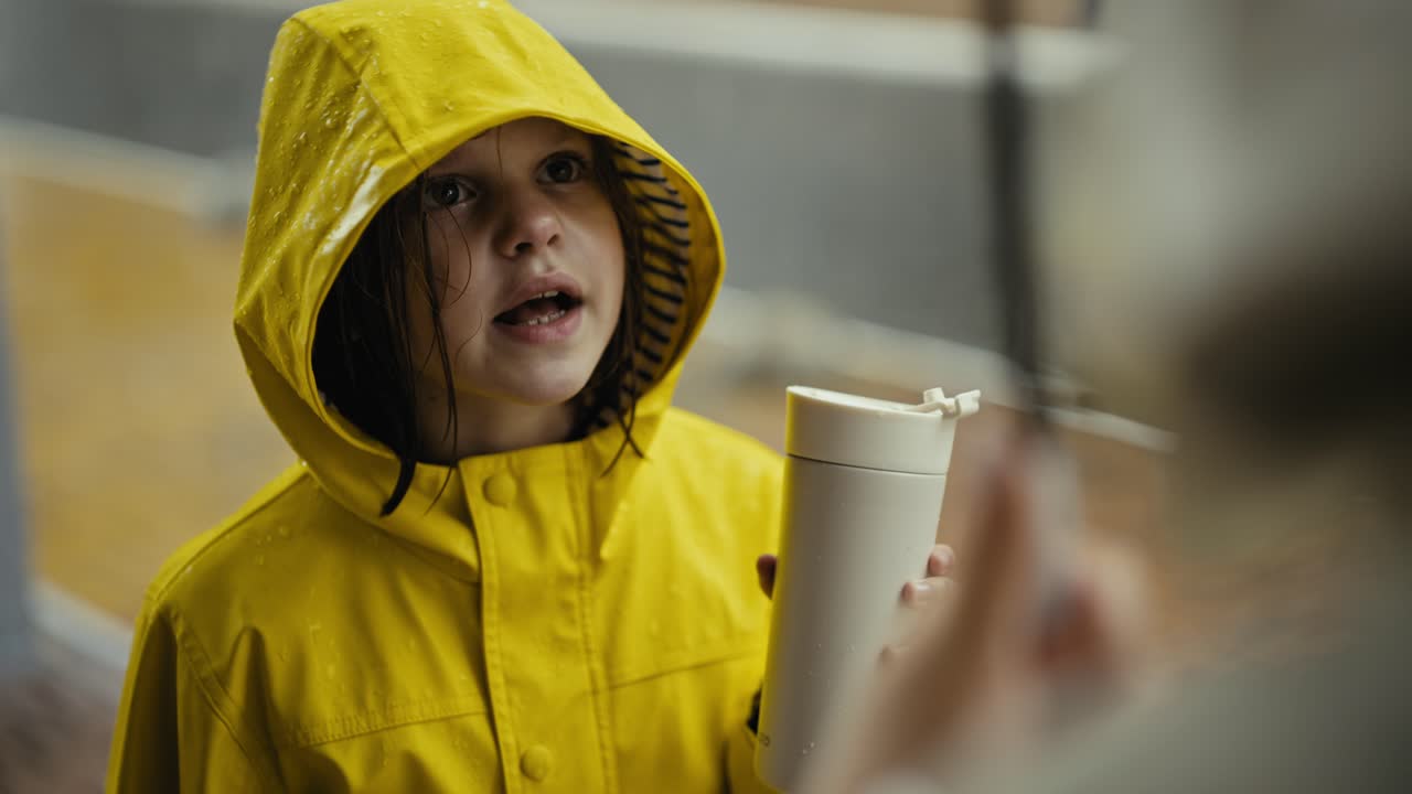 sobre el hombro de cerca de una joven adolescente feliz en una chaqueta amarilla hablando con su madre y bebiendo agua en el parque durante la lluvia