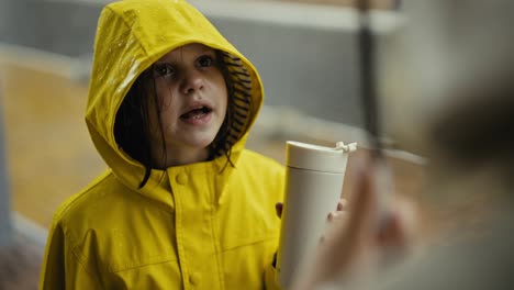 Over-the-shoulder-close-up-of-a-happy-little-teenage-girl-in-a-yellow-jacket-talking-to-her-mom-and-drinking-water-in-the-park-during-the-rain