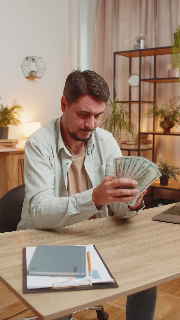 joven caucásico sonriente contando dinero en efectivo usando teléfono inteligente en casa mesa de la oficina en la sala de estar