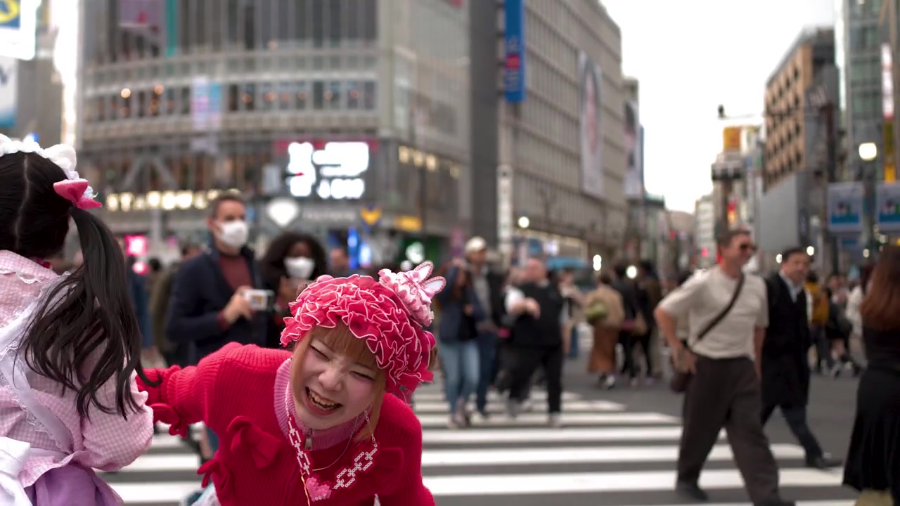 Two women in colorful decora fashion dance and laugh at the busy Shibuya Crossing intersection