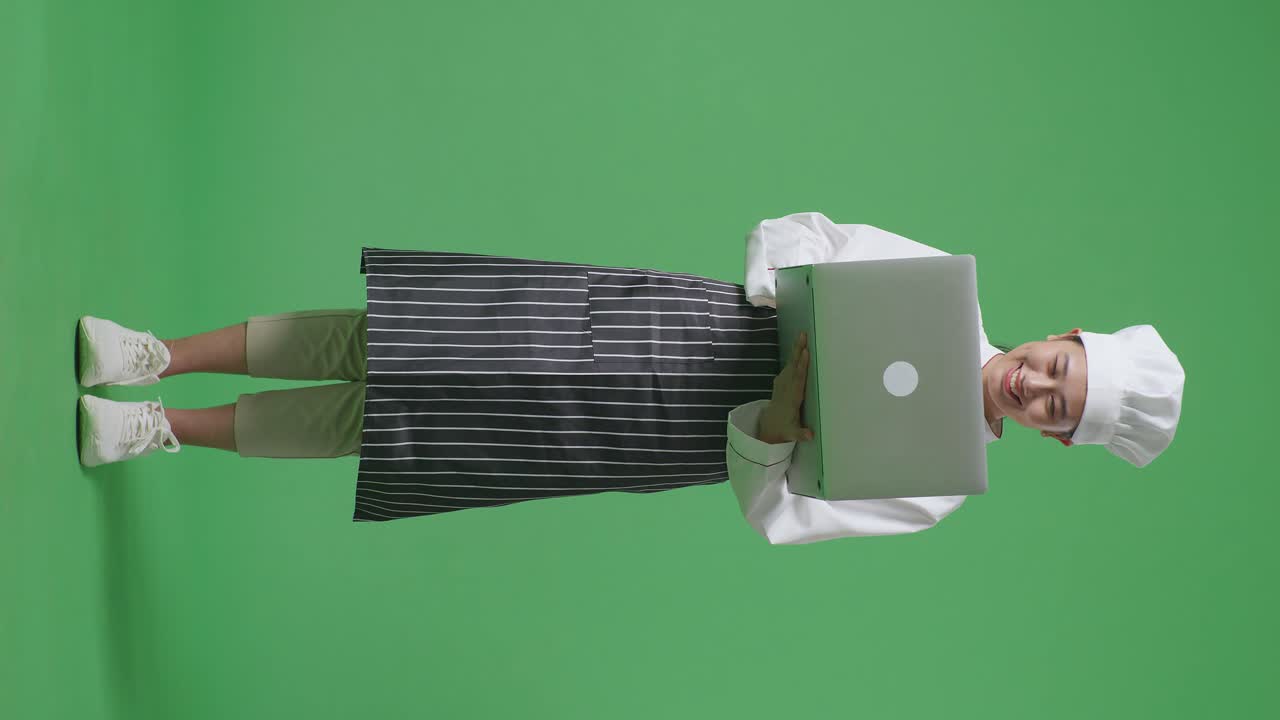 Full Body Of Asian Woman Chef Using A Laptop And Pointing To Side While Standing In The Green Screen Background Studio