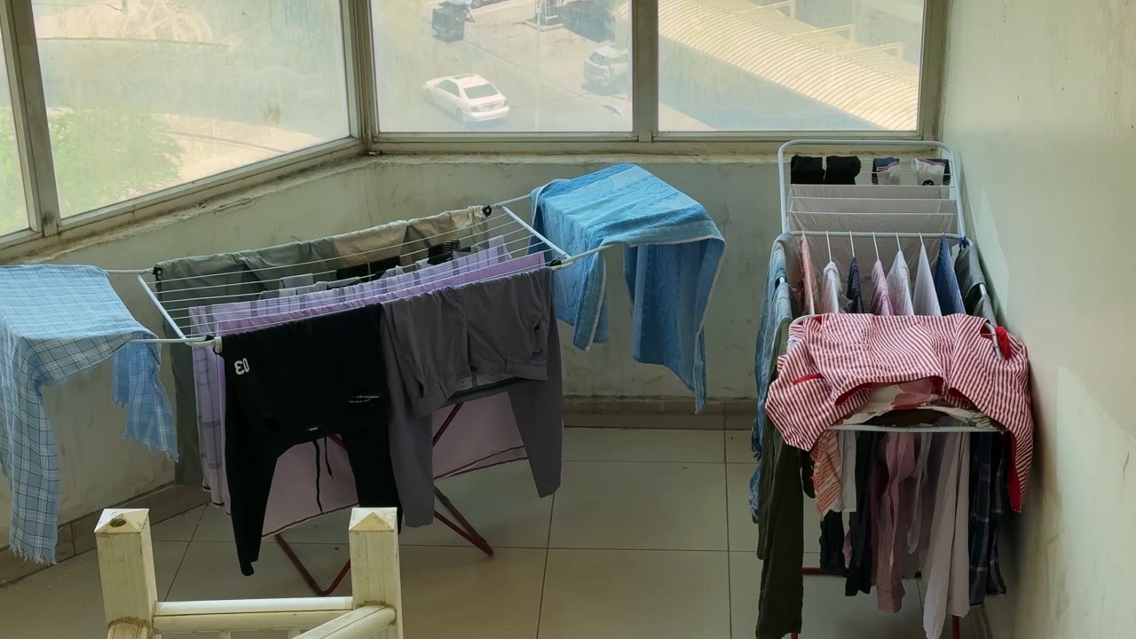 Wet clothes hang on two metal drying racks inside a sunlit enclosed balcony. Daylight streams through large windows overlooking an urban street in this everyday household laundry scene.