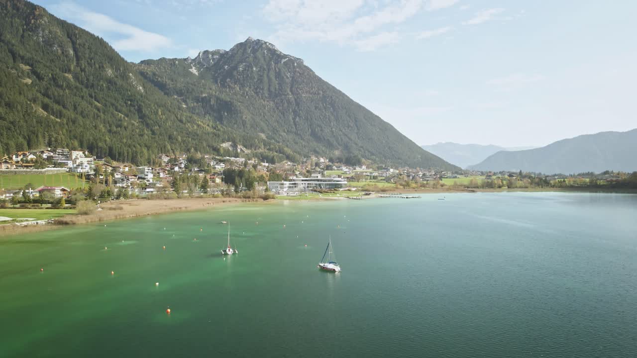 A captivating drone shot at Achensee on a sunny summer day, flying backward to reveal vibrant turquoise water, two boats, and the serene alpine beauty of Austria’s largest lake.