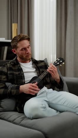 Vertical-video-Happy-blond-man-with-stubble-in-a-plaid-shirt-and-white-T-shirt-sits-on-a-gray-sofa-and-plays-a-black-ukulele-in-a-modern-apartment-during-the-day