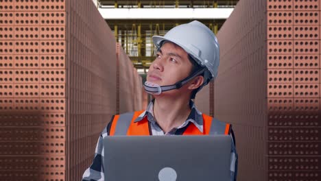 close up of asian male engineer with safety helmet working on a laptop and looking around while standing with red brick packed in stacks are stored