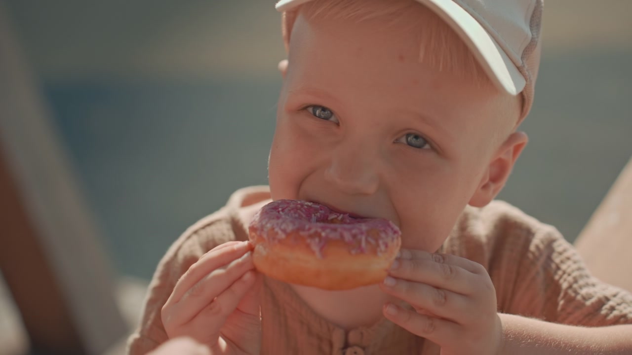 Un niño pequeño recibe y come un donut glaseado rosa de otro niño en un entorno al aire libre. El niño interactúa con la comida mostrando interés mientras se lo acerca a la boca. Luz natural suave