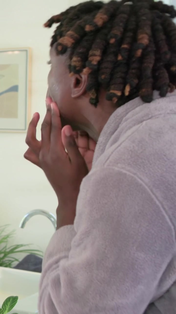 Vertical video: Leaning man with locs in gray robe massaging face at sink, mirror drawing partner