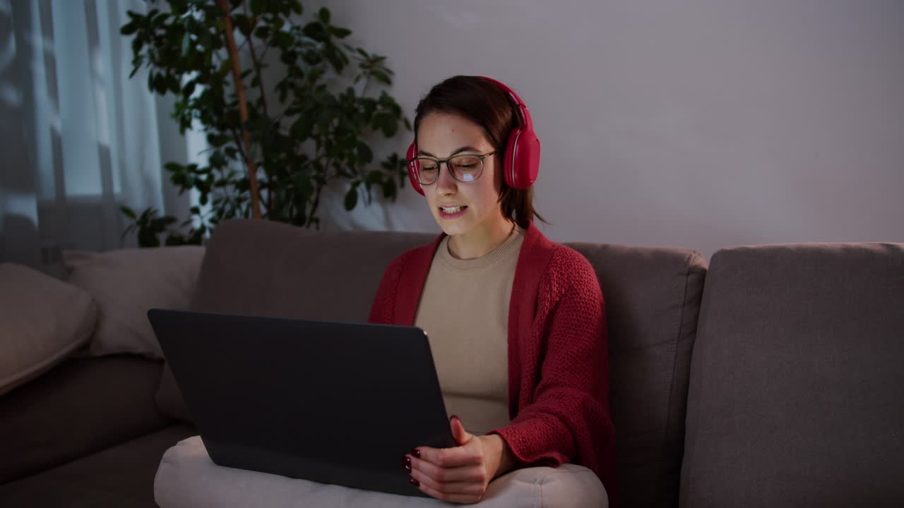 Side view of a confident brunette girl in wireless headphones and glasses sitting on a gray sofa and studying pronunciation in foreign languages using online lessons on gray laptops in a modern apartment