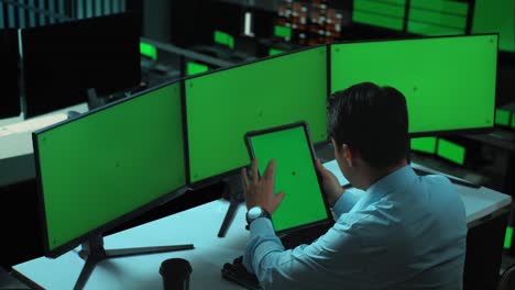 vista lateral de un hombre asiático trabajando con una tableta y varios monitores de computadora con una pantalla verde simulada en la oficina
