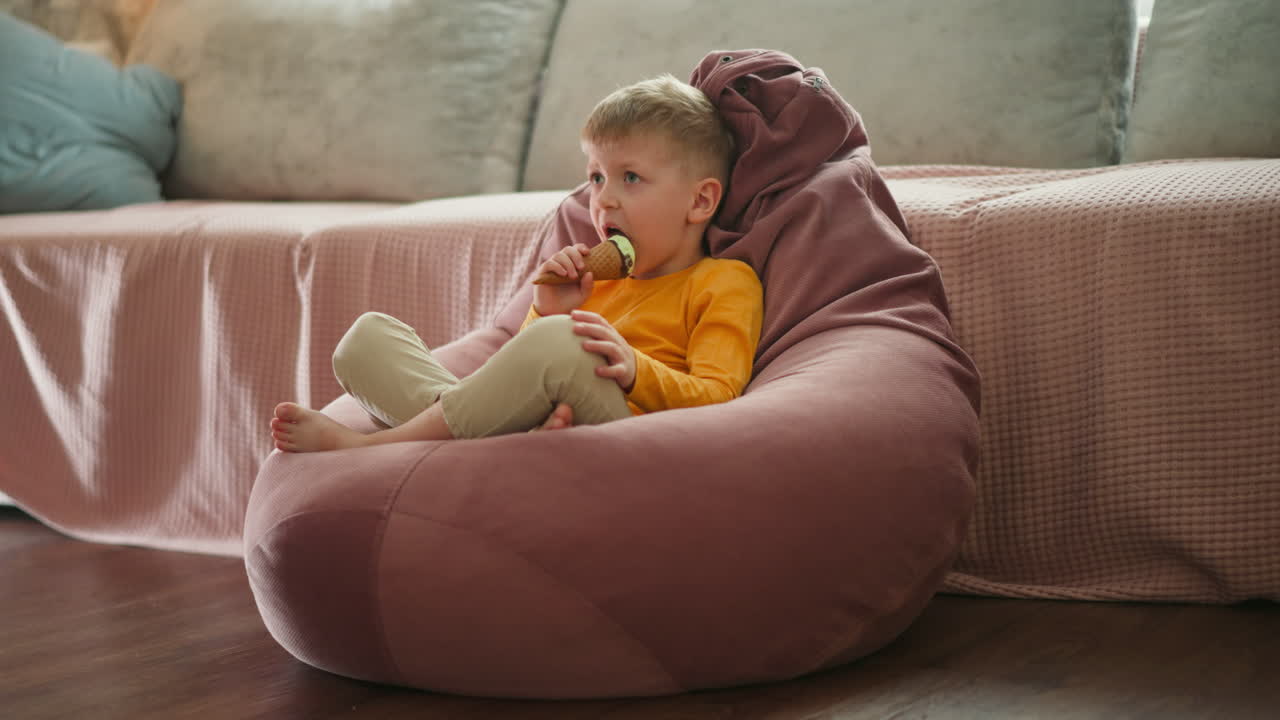 Little boy sitting barefoot on large pink bean bag chair indoors enjoying mint chocolate ice cream cone, looking ahead thoughtfully while relaxing in cozy home setting