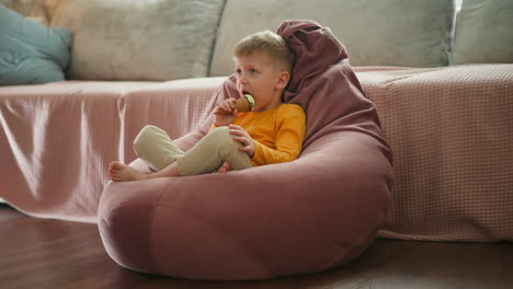 little boy sitting barefoot on large pink bean bag chair indoors enjoying mint chocolate ice cream cone, looking ahead thoughtfully while relaxing in cozy home setting