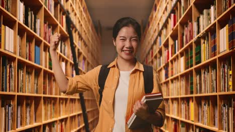 asian woman student with a backpack and some books smiling and flexing her bicep while standing in the library