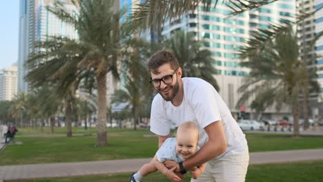 Vater-Spielt-Mit-Söhnen-Im-Sommer-In-Einer-Modernen-Stadt,-Hält-Ihn-Und-Umarmt-Ihn,-Stehend-In-Einem-Weißen-T-Shirt-Und-Shorts