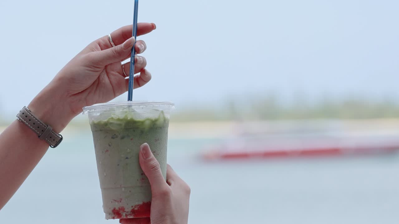 Hands stirring a green iced drink with a straw against a serene beach backdrop, captured in natural daylight