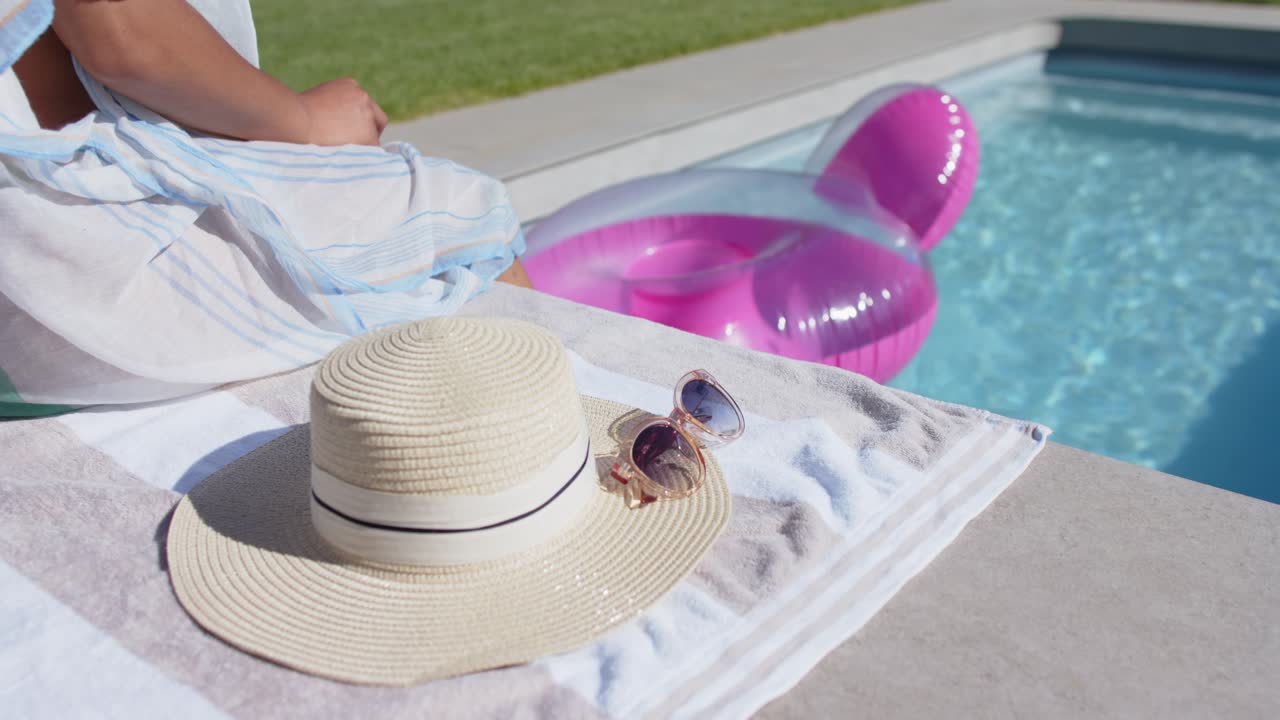 Sun warming African American woman sitting poolside adjusting wrap extending foot onto pink float