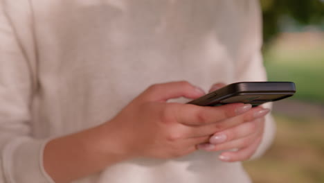 close-up of individual's hand typing on smartphone with polished nails against softly blurred natural greenery background and unclear figures in the surrounding