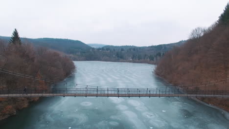 aerial: a man on crutches crosses a wooden cable-stayed bridge over a frozen lake