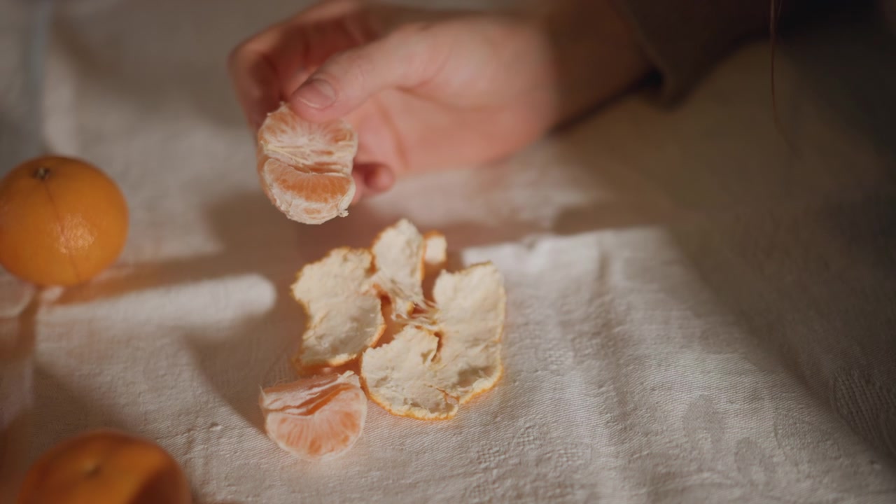 Primo piano di mani caucasiche che sbucciano un mandarino su un morbido asciugamano di lino, luce solare calda che proietta ombre delicate, mandarini sparsi e buccia testurizzata, spicchi succosi rivelati durante un lento rituale di spuntino casalingo, accogliente.