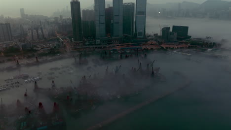 Wide-angle-slow-revealing-aerial-shot-of-fog-covered-West-Kowloon-with-loading-crane-barges-docked-in-Typhoon-shelter,-Hong-Kong,-at-dawn