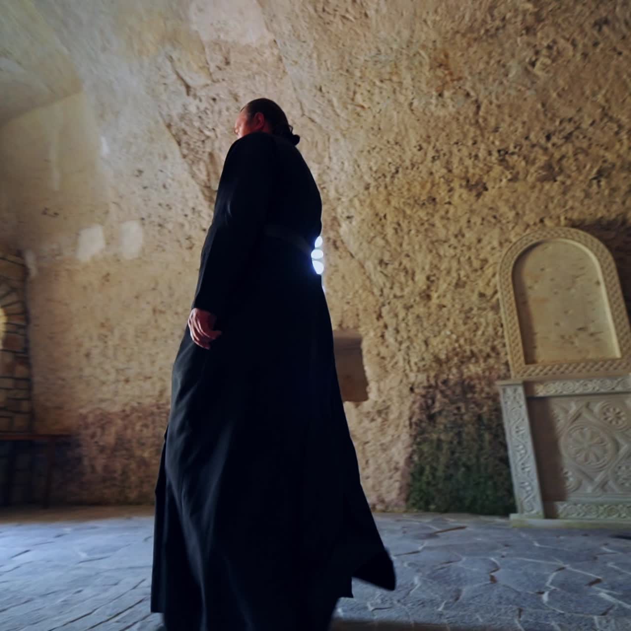 Orthodox priest in the spacious chapel cut in the rock. Monk walking to the doorway of the monastery with bright daylight. Low angle view