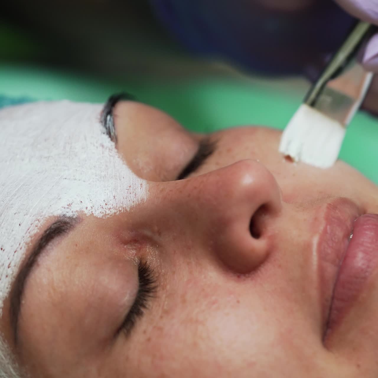 Applying white face mask on female face. Woman in blue protective cap having facial care at beautician's. Extreme close up