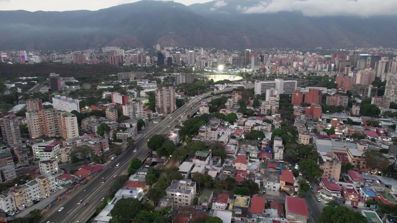 Drone che vola all'indietro sopra Los Chaguaramos, mostrando l'autostrada di Caracas e il paesaggio urbano al tramonto.