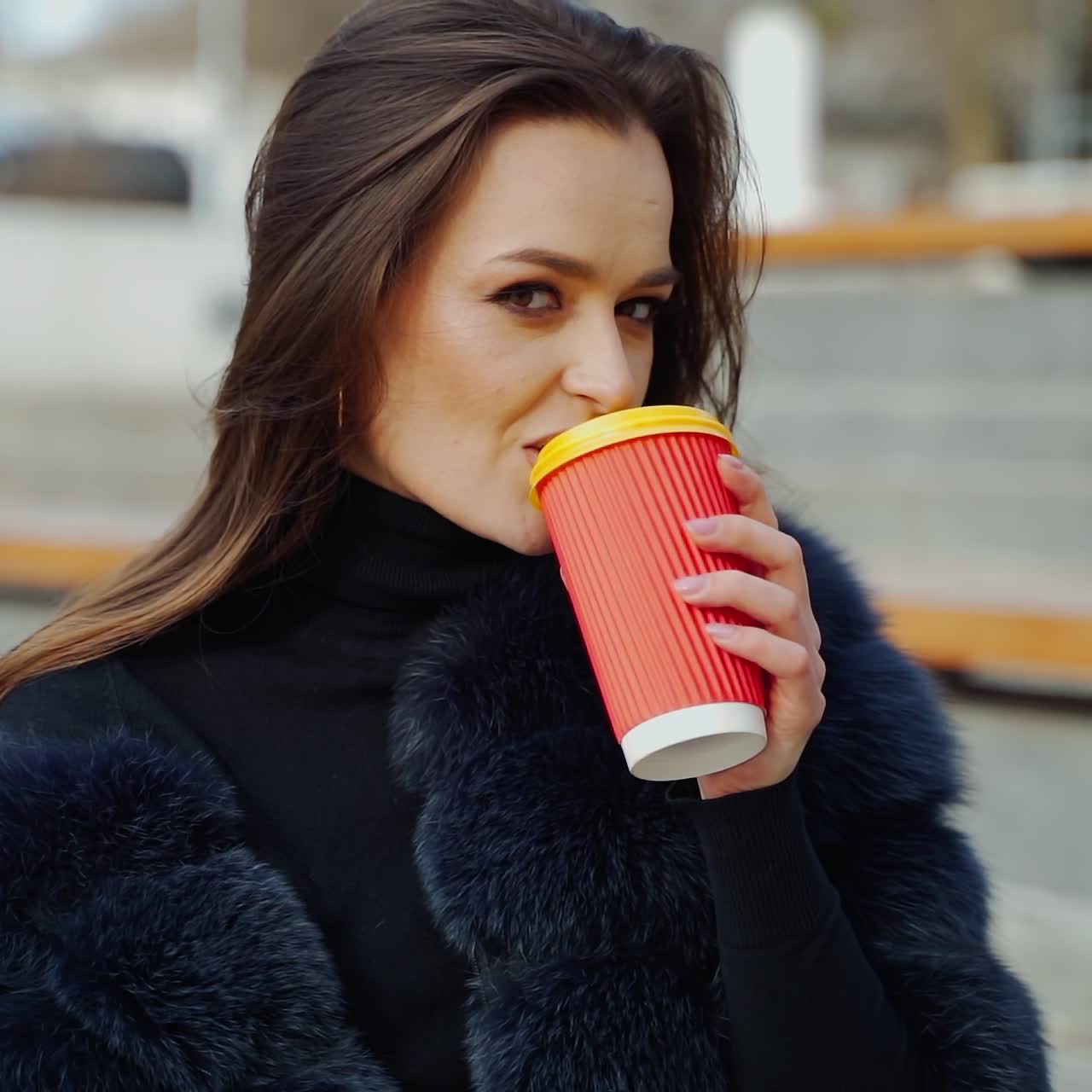 Female hands hold a cup of beverage outdoors. Beautiful woman in a fur coat sitting in the park and drinking from plastic glass. Slow motion.