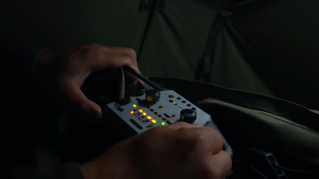 Close-up of hands operating a compact electronic control panel with glowing buttons, knobs, and indicator lights in a dim indoor setting with cables