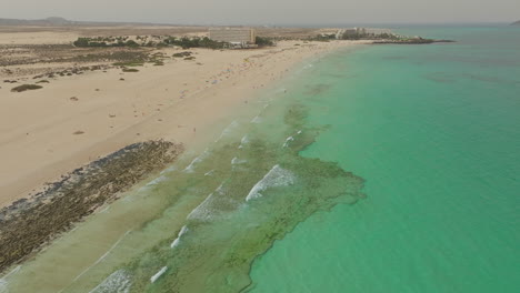 aerial-view-in-orbit-over-the-shore-of-Corralejo-beach-and-its-turquoise-waters,-on-the-island-of-Fuerteventura-on-a-sunny-day