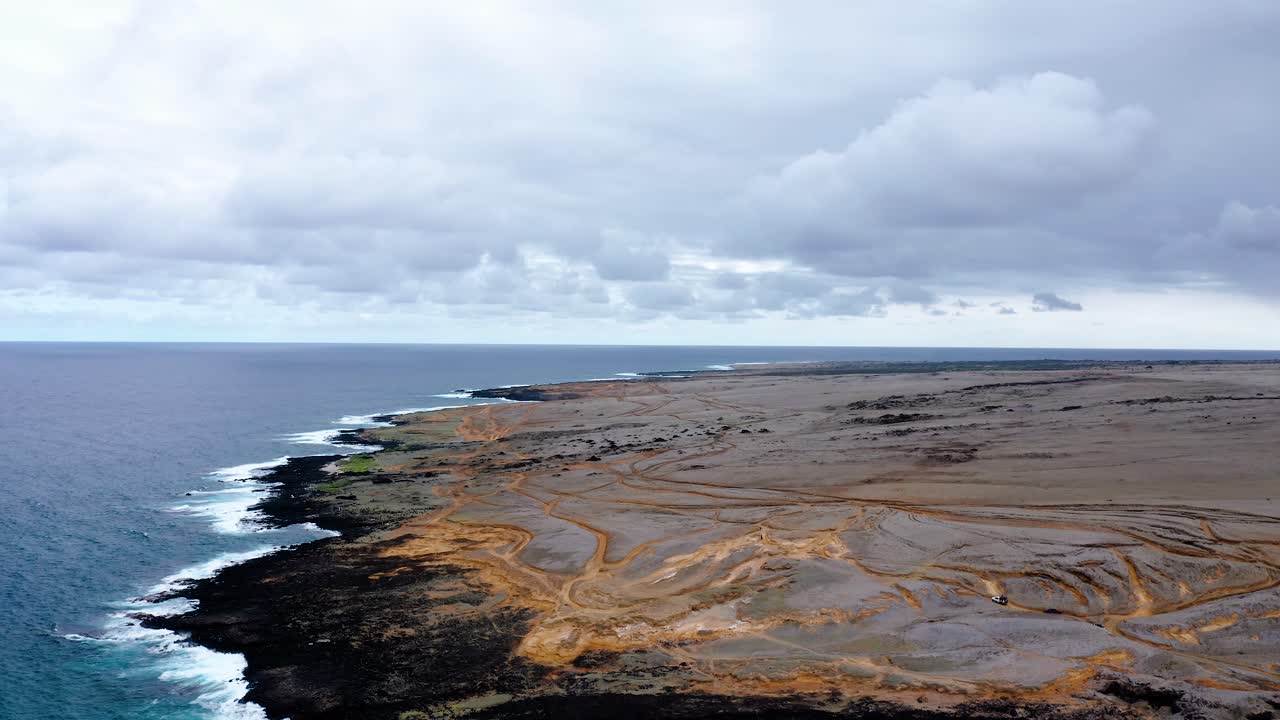 Expansive view of a remote volcanic coastline with rugged black rocks, winding dirt trails, and a moody sky above the ocean horizon.